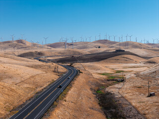 Fototapeta premium Aerial view shows vehicles driving along a winding asphalt highway through arid rolling hills, leading toward a large wind farm with spinning turbines in California.