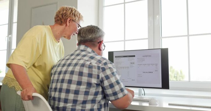 Senior couple reviews electricity bill and online payment. They compare a paper statement and point at charges on a desktop computer in home office. Digital household budgeting made easier.