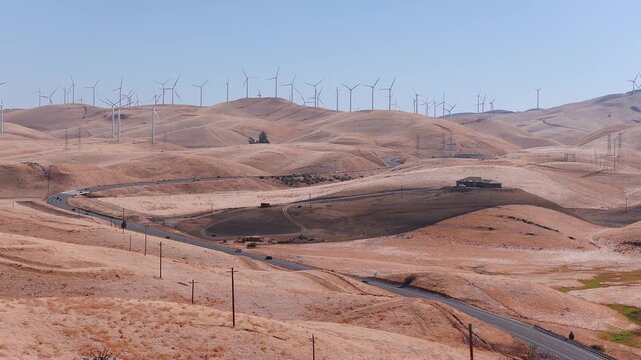 Aerial view shows modern wind turbines spinning across dry rolling hills beside a highway in Altamont Pass, California, generating sustainable renewable power.