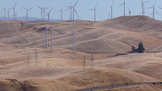 An aerial sequence captures numerous modern wind turbines spinning across dry golden rolling hills at Altamont Pass in California, generating clean renewable electric power under daylight.