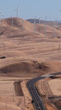 Aerial view shows modern wind turbines spinning across arid rolling hills in California beside a curving multi lane highway carrying traffic through Altamont Pass on a sunny day.