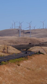 Cars and trucks drive along a winding rural highway past dry grassy hills and spinning wind turbines at Altamont Pass Wind Farm in California under clear sunny skies.