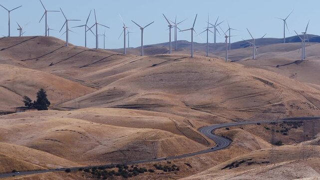 Aerial shot captures numerous wind turbines spinning along the dry golden hills of Altamont Pass in California, with a winding road curving below through the valley.