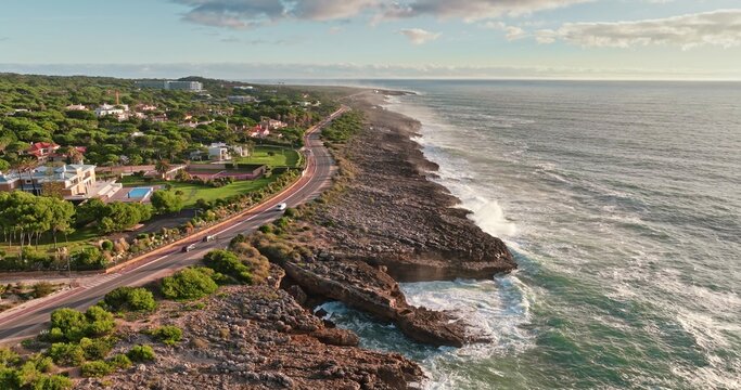 Aerial view of a coastal road winding along rocky cliffs and the Atlantic Ocean, bordered by lush green trees and residential villas in Lisbon, Lisbon, Portugal.
