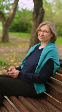 Vertical video of beautiful smiling mature blond woman using her phone for texting messages in park
