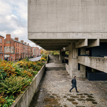 A pedestrian walks past a massive modern brutalist concrete building contrasting with traditional red brick row houses on a cloudy day