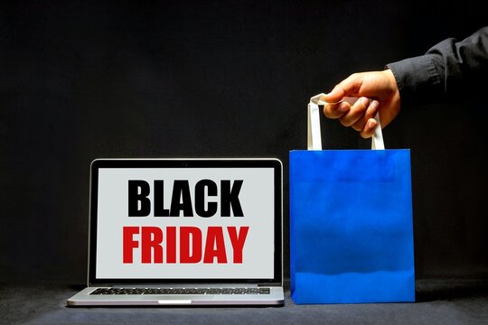 Laptop showing Black Friday sign on screen next to hand holding blue paper shopping bag