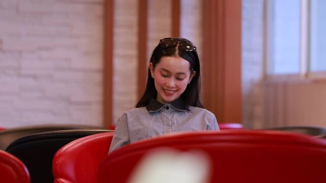 Young Asian woman studies reading book anf Siting on red chair in quiet library. Books line tall shelves behind. she holds book, calm concentration. Ideal for educational or study-themed projects