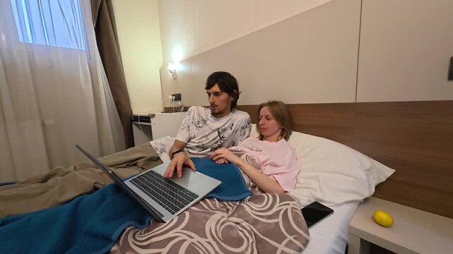 Couple sits close together in bed with laptop on their laps, sharing relaxed moment at home. Young man and woman look toward screen under warm room lighting, seated among pillows and bedding.