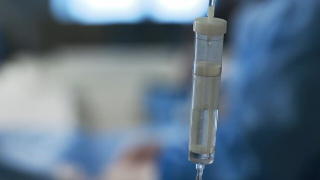 Intravenous infusion drip chamber in focus against blurred operating room with surgeons, medical treatment and patient care concept