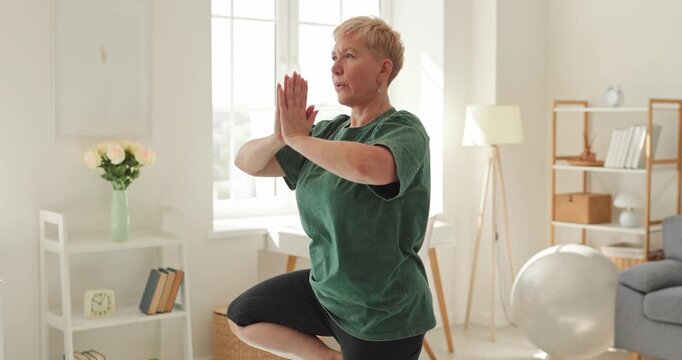Senior woman holds yoga tree pose balance. Footage shows her raise arms and join palms while maintaining a steady one leg stance at home. Mindful movement supports healthy independent living.