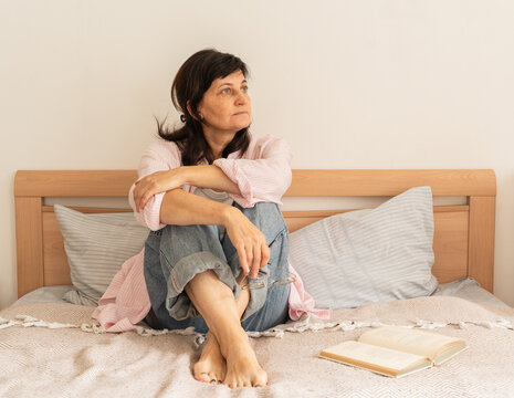 Mature woman in jeans and pink shirt sits on bed, arms wrapped around her knees, looking pensive and gazing off to side, book and glasses lie nearby. Measured pace of life, mindful leisure