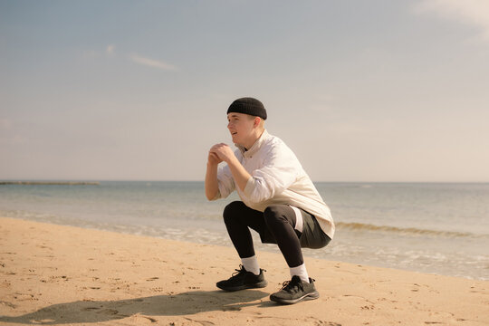 Young active man doing squats for warming up on sandy beach before outdoor workout or running