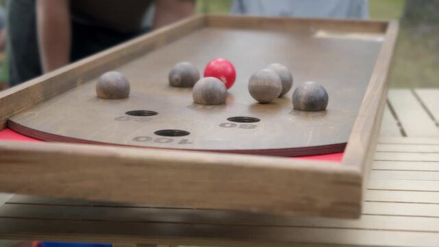 Young child tilts rustic wooden board trying to guide rolling balls into scoring holes. Game pieces roll past targets as kid misses making successful shot during outdoor leisure activity.