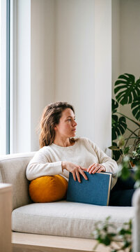 Pensive young woman relaxing on a comfortable sofa holding a book and looking out the window in a modern bright living room