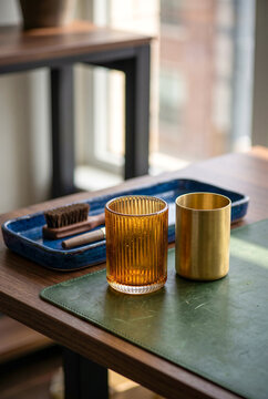 Amber ribbed glass cup and brass tumbler placed on a green leather desk mat next to a blue ceramic organizer tray