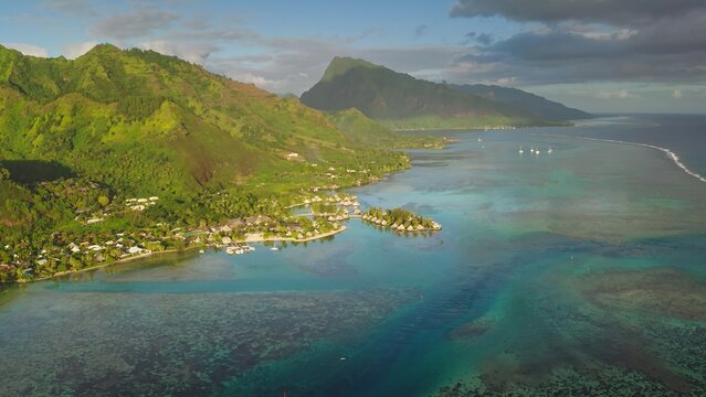 Aerial view of the lush green mountains and turquoise lagoon of Moorea, featuring overwater bungalows and boats in the clear water, Moorea-Maiao, Windward Islands, French Polynesia.