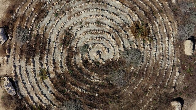 Aerial view of a stone spiral labyrinth set in dry, rocky terrain with sparse vegetation, Cyprus.