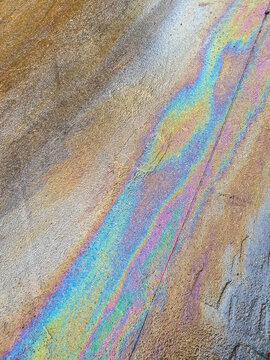 A vibrant iridescent rainbow sheen from an oil slick spreads across a rough, coarse textured asphalt ground surface, brilliantly illuminated by bright natural daylight