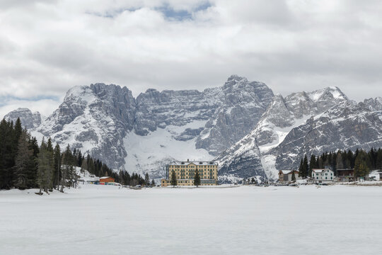 Snow covers the landscape around Lake Misurina in the Dolomites of Italy. Mountains rise behind a building set in the winter scene.
