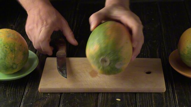 Hands place a large, spotted melon on a wooden cutting board and begin paring off the skin with a small knife, while other fruits lie on plates nearby