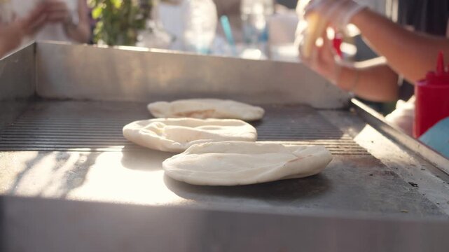 Global street food: diverse cultural flatbreads, such as pita or bazlama, are baking. A professional chef prepares fresh traditional dough on hot surface near red sauce dispenser and background crowd.