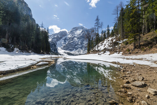Snow covers the ground and trees around Lake Braies in the Dolomites at winter. Mountains reflect in the water under a clear sky.