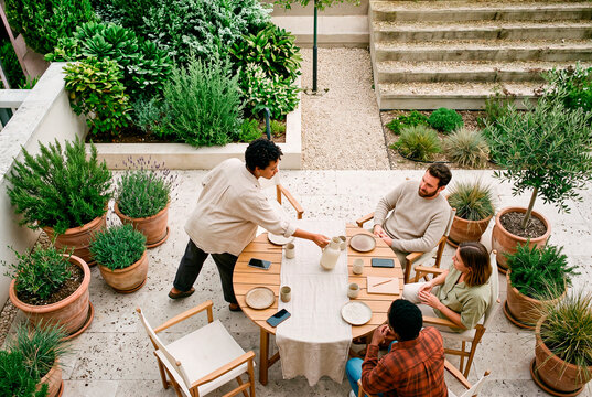 Diverse group of friends gathering around a wooden dining table on a sunny outdoor patio surrounded by plants