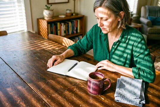Thoughtful mature woman sitting at wooden table with open notebook and coffee mug in sunny living room