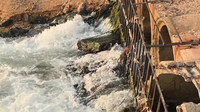 Industrial wastewater discharging through concrete culvert outlet with rusted metal grate into turbulent rocky channel