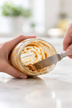 Hand scraping the last of the peanut butter from a nearly empty glass jar with a knife