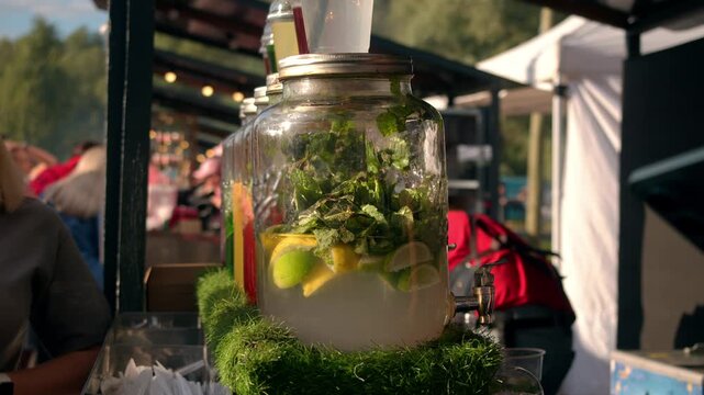 Close up shows large glass dispenser full of fresh citrus lemonade. Refreshing summer beverage features mint leaves and sliced lemons resting on artificial grass at sunny outdoor food market.