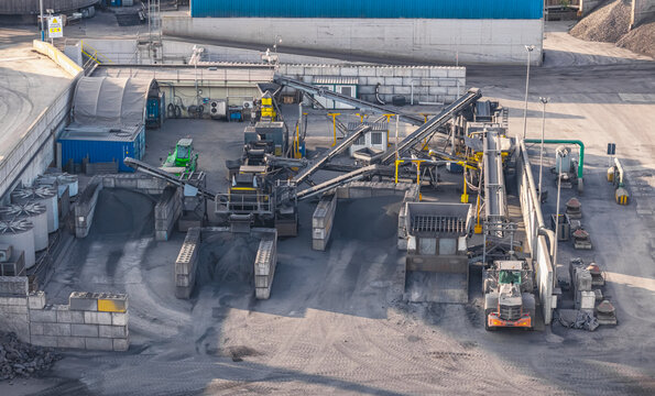 Aerial view of industrial material handling and conveyor system processing dark raw materials inside a large heavy manufacturing and metal production facility in Brescia, Lombardy, Italy.