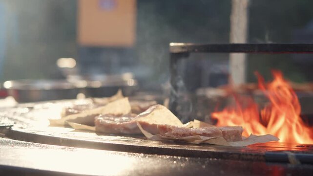 Gloved chef hand places raw meat patty on brown craft paper over hot metal grill. Bright orange flames rise from barbecue station during busy outdoor street food festival.
