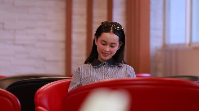 Young Asian woman studies reading book anf Siting on red chair in quiet library. Books line tall shelves behind. she holds book, calm concentration. Ideal for educational or study-themed projects