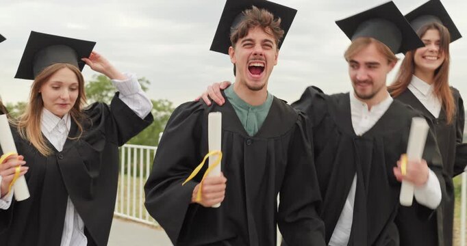 Graduate friends celebrate outdoors with diplomas and smiles. Young students in caps and gowns laugh and dance toward camera during ceremony. A very joyful moment of shared achievement.