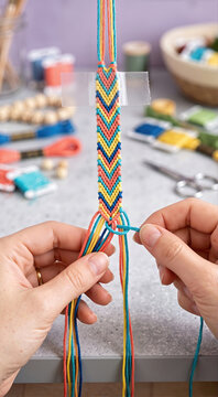 Vertical photo of hands weaving chevron pattern friendship bracelet. DIY handmade jewelry process on grey craft desk. Creative hobby with colorful threads