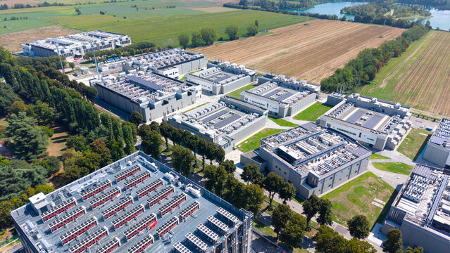 Aerial view of a modern data center campus featuring multiple server buildings with rooftop cooling units, surrounded by green trees and agricultural fields in Milan, Lombardy, Italy.