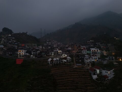 Aerial view of the Dieng village nestled in the mountainous landscape with illuminated houses and terraced fields under a cloudy sky in Kejajar, Central Java, Indonesia.