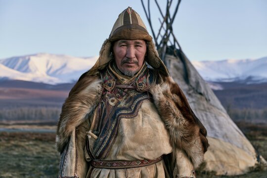 Indigenous Siberian man in fur trimmed traditional clothing and pointed hat standing before a conical tent in a snowy mountain landscape, portrait