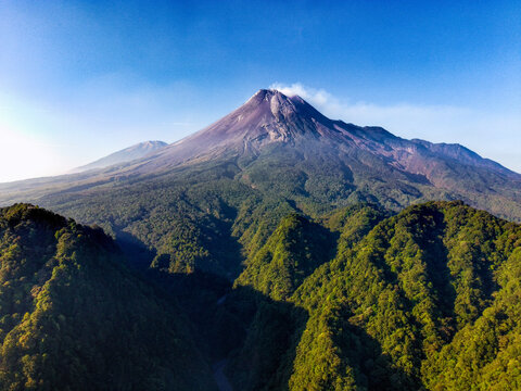 Aerial view of Mount Merapi with lush green forest slopes, volcanic terrain, and clear blue sky in Kecamatan Pakem, Daerah Istimewa Yogya, Indonesia.
