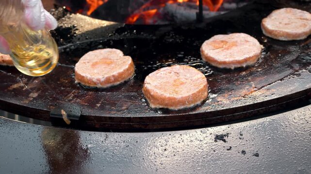 Generous stream of golden sunflower oil pours onto hot metal flat grill. Raw meat burger patties sizzle on oiled surface near open fire at busy street food market.