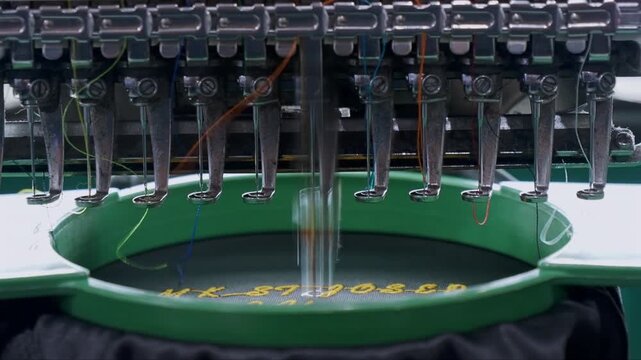 A detailed industrial shot of a multi-head embroidery machine. Multiple needles with colorful threads are poised above a blue fabric taut in an embroidery hoop.