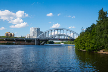 Horizontal view of a picturesque white arch bridge spanning a blue river, with a small cruise ship passing beneath it. Excellent for travel themes, cityscapes, and peaceful summer wallpapers. © Margarita