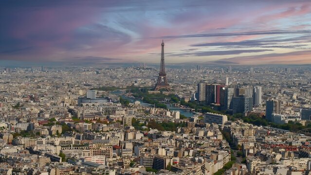 Aerial view of the Eiffel Tower rising above the dense urban landscape of Paris, featuring the winding Seine River and historic rooftops under a dramatic sunset sky in Paris, Ile-de-France, France.