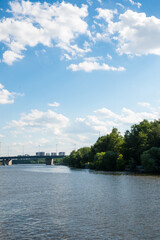 Vertical landscape of a wide river flowing past a green forest bank, a distant bridge, and a bright blue sky with fluffy clouds. Great for nature wallpapers, travel themes, and peaceful backgrounds. © Margarita