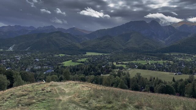 Panoramic view of a mountain town situated in a green valley below majestic rugged peaks under dramatic moody cloudy sky. Zakopane city view