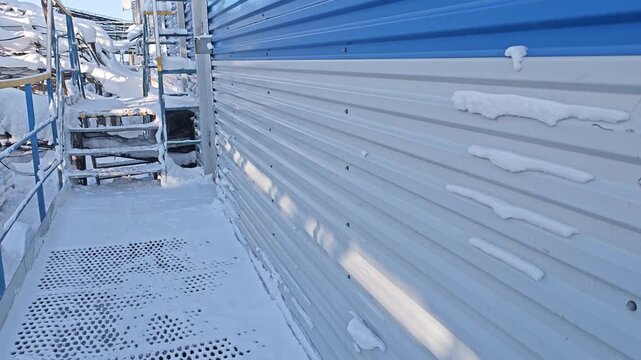 Winter maintenance on snow covered metal structure continues along narrow walkway. Blue handrails and ladder access line corrugated panels, and frozen platform suggests harsh outdoor conditions.