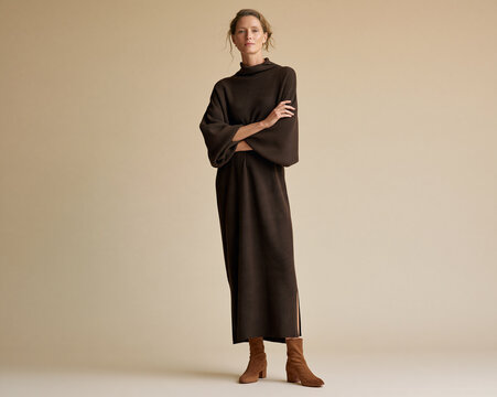 Sophisticated woman in a loose chocolate-brown knit maxi dress with wide sleeves and suede ankle boots poses against a warm neutral studio background.
