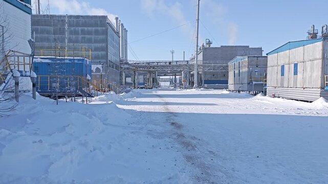 Snowy service road cuts through industrial utility access beside concrete buildings and pipework. This cold, empty facility has stark, orderly look, with faint steam lifting into blue winter sky.